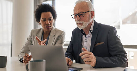 Female,Executive,Discussing,With,Senior,Professional,Over,Laptop.,Business,Colleagues